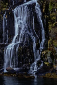 Waterval die over rotsen stroomt met mos en gras, mondend in een kleine plas water.