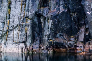 Rotswand met verticale scheuren en gele mosplekken, weerspiegeld in water.