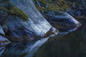 Rotsachtige oever met mos en begroeiing, reflecties in kalm water.