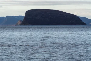 Groot rotsachtig eiland omringd door water met besneeuwde bergen op de achtergrond onder bewolkte lucht.