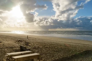 Leeg zandstrand met houten zitbanken onder bewolkte hemel en zonlicht aan kustlijn.