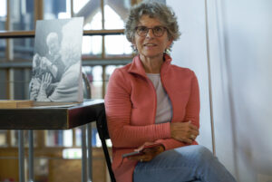 Vrouw met bril in oranje jas zittend bij tafel met zwart-wit foto van breiende mensen.