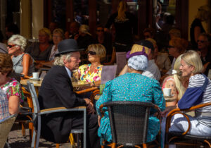 Druk terras cafe met oudere mensen in zonnige sfeer, converseren aan ronde tafels.