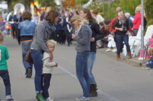 Straatfeest met volwassenen en kinderen in gesprek, midden twee vrouwen en kind, kramen op achtergrond.