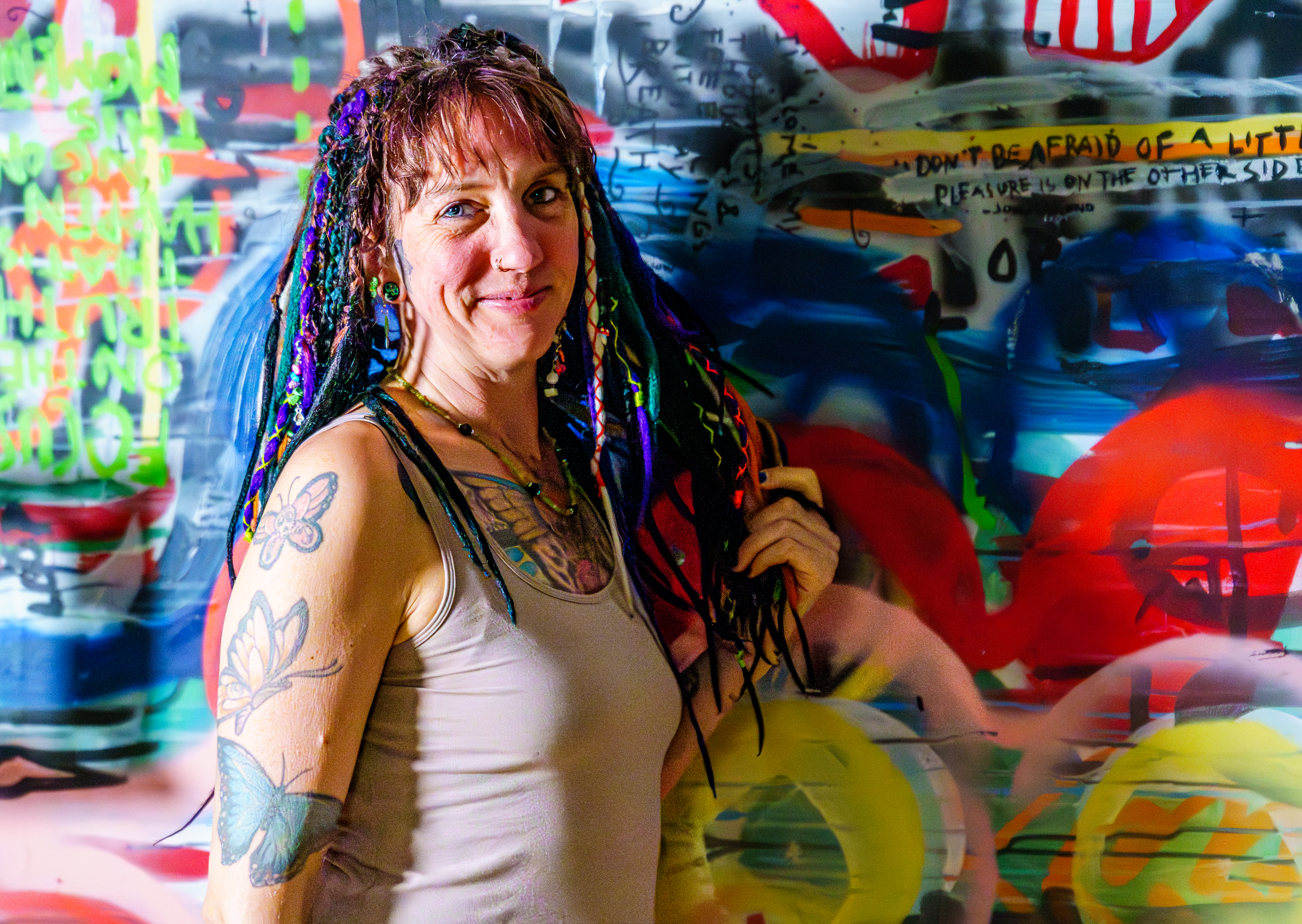 Vrouw met kleurrijke vlechten en tattoos staand voor een kleurrige graffiti-achtergrond, glimlachend naar camera.