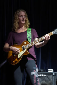 Een vrouw speelt elektrisch gitaar op podium, blik naar beneden, groene snakelaarband, paarse top.