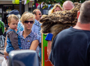 Een vrouw met zonnebril draagt een jongen terwijl ze tussen een menigte op een straatfestival staat.