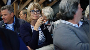 Vrouw met bril houdt glas wijn omhoog tijdens bijeenkomst, omringd door pratende mensen.